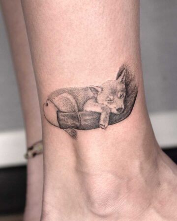 A delicate fine line dog tattoo of a sleeping puppy curled up in a bed on an ankle. This beautiful animal tattoo is a sweet memorial, perfect for pet lovers.