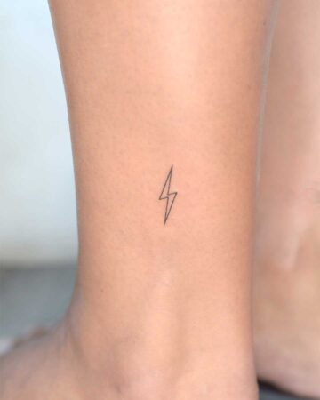 A small, minimalist lightning bolt tattoo on a person's ankle. The black outline of the bolt is simple and elegant, a common design for small tattoos, perfectly placed on the skin. This minimalist tattoo is an example of a popular ankle tattoo choice.