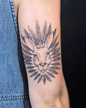 A unique tattoo of a kangaroo head surrounded by feather-like designs, inked on an upper arm. The kangaroo has a mole on its nose and striking blue eyes, set against a denim-clad shoulder.