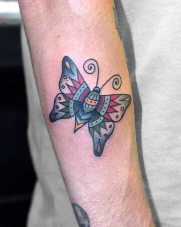 A vibrant American traditional butterfly tattoo on a forearm. The design features bold black outlines, red, blue, and green details, and is a classic example of tattoo art. This butterfly tattoo is a striking example of traditional tattoo style.