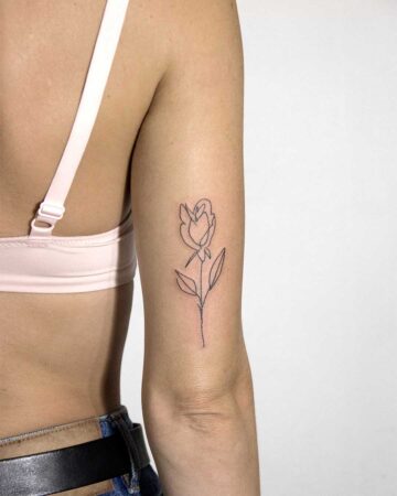 A woman's arm showcases a delicate, minimalist flower tattoo. The fine-line design depicts a rose with a slender stem and leaves, a perfect example of a small tattoo. Done by tattoo artists in our fine line tattoo studio parlour shop Canggu, Bali