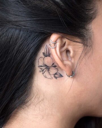 A woman's ear with a delicate behind-the-ear tattoo of a flower and leaves. The black and white design is a small tattoo, showcasing elegant lines and detail. The ear has multiple piercings with silver hoops.