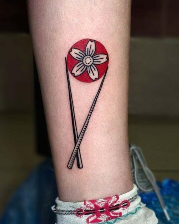 Tattoo of crossed chopsticks holding a white flower with a red circle behind it, on a person's lower arm.