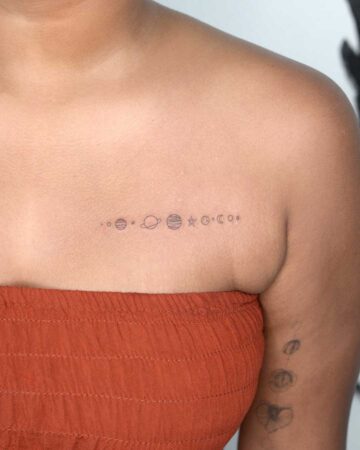 Chest tattoo of the planets in our solar system in minimalist style on a woman with an orange strapless top. A second tattoo of flowers is visible on her arm.