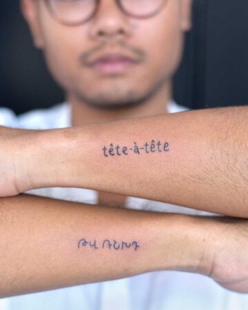 Close-up of a person's forearms displaying tattoos: "tête-à-tête" in French and a word in Armenian script. Focus on unique tattoo designs.