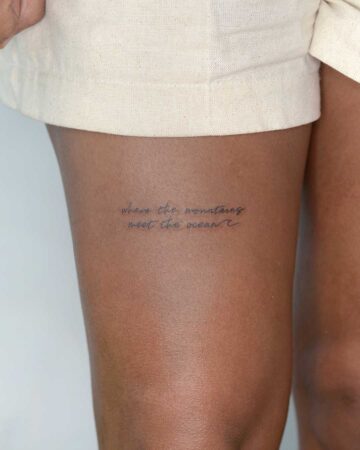Close-up of a thigh with a delicate script tattoo that reads: "where the mountains meet the ocean". The tattoo is in black ink, suggesting adventure and travel.