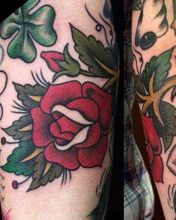 Close-up of a vibrant traditional tattoo featuring a red rose with white highlights, surrounded by green leaves and a shamrock. Classic tattoo art style.