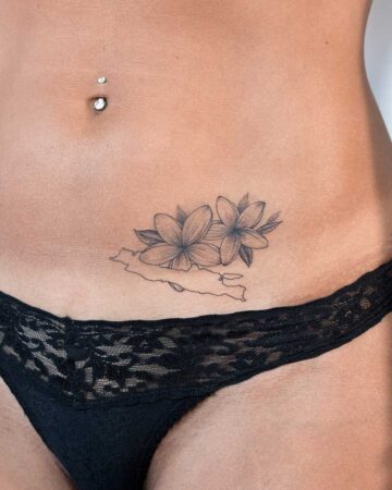 Close-up of a woman's lower abdomen featuring a delicate tattoo of two plumeria flowers above an outline of an island or landmass, positioned above black lace underwear and a navel piercing.