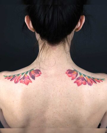 Back view of a woman with dark hair in a bun, showcasing vibrant floral tattoos on her upper back and shoulders. The tattoos feature red and orange flowers with green stems, creating a striking contrast against her skin.