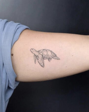 Delicate black ink sea turtle tattoo on an arm, featuring a minimalist design. The small tattoo captures the turtle's form with detailed lines and shading.