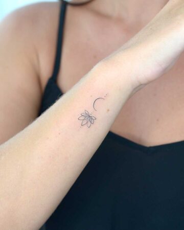 Delicate minimalist tattoo of a lotus flower and crescent moon on a woman's wrist. The fine-line tattoo design is a popular choice for a wrist tattoo, showcasing a lotus tattoo and moon tattoo.
