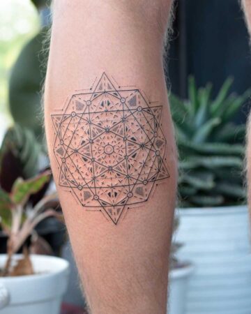 Intricate geometric tattoo on a calf, featuring a detailed pattern of lines, dots, and triangles. The design is black ink on fair skin, showcased with a blurred background of potted plants.