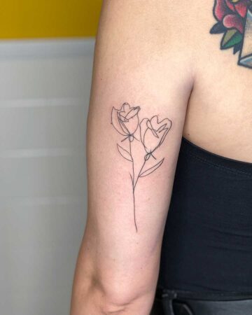 Minimalist fine-line tattoo of two delicate tulip flowers on a woman's upper arm. A vibrant, colorful tattoo peeks out from her shoulder, adding a pop of contrast to the simple floral design.