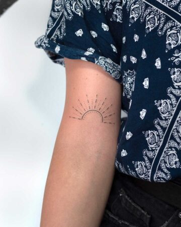 Minimalist sun tattoo on a woman's inner arm, featuring a semi-circle with radiating lines, against a backdrop of a blue patterned shirt and jeans.