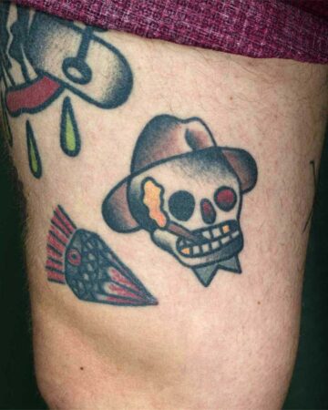 Traditional tattoo art on skin: a skull in a fedora smoking a cigar, plus a stylized teardrop and a red-finned fish. Classic tattoo flash design.