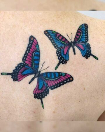 Two vibrant butterfly tattoos on a person's shoulder. The butterflies feature intricate designs with pink, blue, and black ink, showcasing a beautiful example of colorful body art and butterfly tattoo ideas.