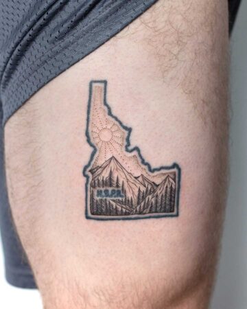 Idaho tattoo featuring a mountain range and pine trees within the state's outline. The design includes a sunburst and the letters "H.S.P.R." inside the Idaho shape. A travel-inspired design for Idaho's natural beauty. Done by tattoo artists in our fine line tattoo studio parlour shop Canggu, Bali