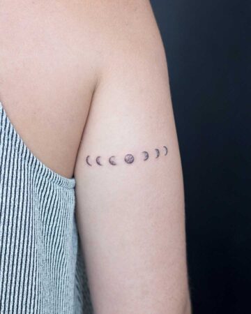 A person's arm displays a minimalist moon phase tattoo, a series of small, delicate crescent and circular shapes representing the moon's cycle. The arm is positioned against a dark background, highlighting the simple, elegant design, a perfect example of a small moon phase tattoo.