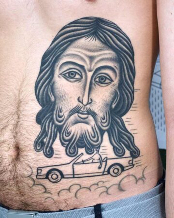 A large, detailed tattoo of Jesus' face dominates the abdomen, with flowing hair and beard. Below, a small figure drives a convertible car amidst stylized clouds. The tattoo is a unique blend of religious art and modern imagery, showcasing a creative tattoo design. Done by tattoo artists in our fine line tattoo studio parlour shop Canggu, Bali