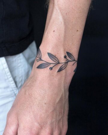 Close-up of a wrist adorned with a delicate leaf tattoo. The minimalist design features a thin, black line drawing of leaves and berries, forming a band around the wrist. A small, elegant wrist tattoo. Done by tattoo artists in our fine line tattoo studio parlour shop Canggu, Bali