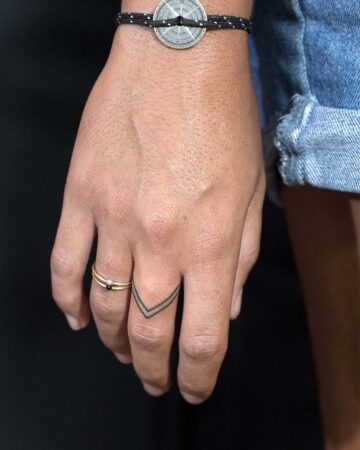 Close-up of a hand showcasing a minimalist finger tattoo, two thin black lines forming a V shape on the index finger. A delicate gold ring with a black stone sits on the ring finger, complemented by a compass bracelet. The hand is against a black background, highlighting the small tattoos. Done by tattoo artists in our fine line tattoo studio parlour shop Canggu, Bali