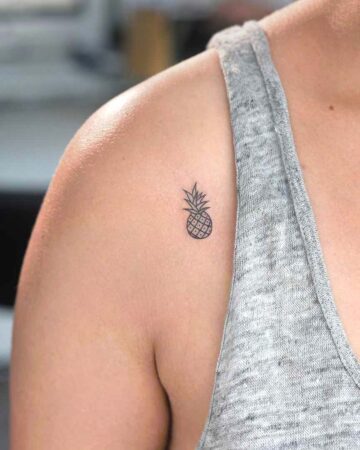 A small, minimalist pineapple tattoo on a woman's shoulder. The delicate black outline showcases the fruit's textured skin and spiky crown, a popular design for small tattoos.