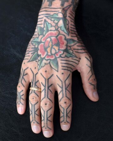 Hand tattoo featuring a traditional rose design on the back of the hand and geometric patterns on the fingers. Visible gold wedding band adds a personal touch.