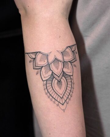 Close-up of a delicate, black ink mandala tattoo on a fair-skinned forearm. The design features intricate dotwork shading and geometric patterns, creating a striking ornamental effect.