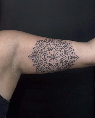 Close-up of a geometric mandala tattoo in black ink on a person's upper arm. The design features symmetrical floral and swirling patterns.
