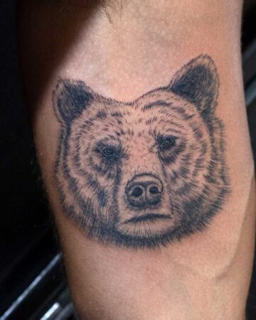 Detailed black ink tattoo of a bear's head on a person's arm. The bear has a somber expression with realistic fur texture and shading.