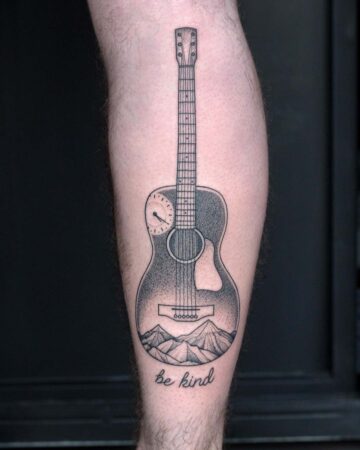 Detailed black ink tattoo of an acoustic guitar featuring mountain range imagery at the base and a clock face, with the text "be kind" beneath.