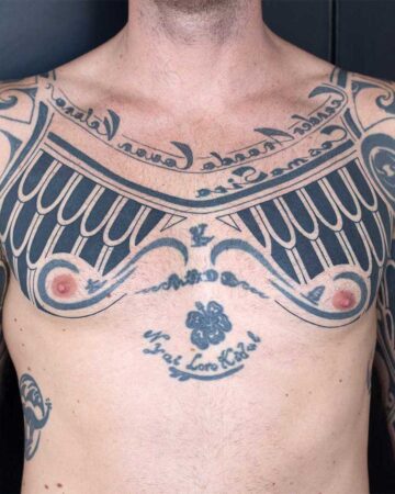 Man's chest with extensive tribal-inspired black ink tattoo art covering the upper chest and sternum. The design incorporates geometric patterns, decorative swirls, and script. Lower chest features a flower and text "Nyat Loro Kidul.