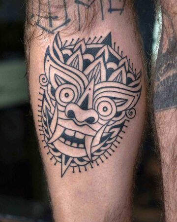 Black ink tattoo on a hairy calf featuring a stylized, symmetrical demon mask with large eyes, sharp teeth, and intricate linework, surrounded by a decorative border.