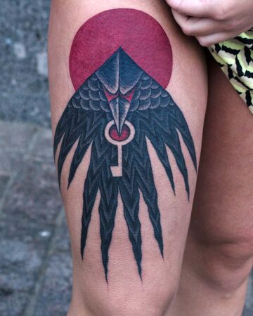 Thigh tattoo featuring a stylized black raven with outstretched wings, holding a silver key against a red circular background. Geometric design.