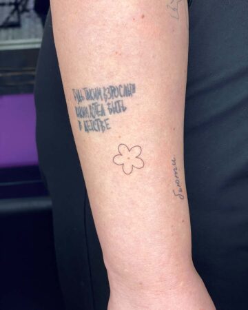 A person's arm displays several tattoos, including a small, minimalist flower tattoo. Above it, faded Russian text reads, "Be the adult you wanted to be as a child." The word "Бьюти" (Beauty) is written vertically on the arm. Done by tattoo artists in our fine line tattoo studio parlour shop Canggu, Bali
