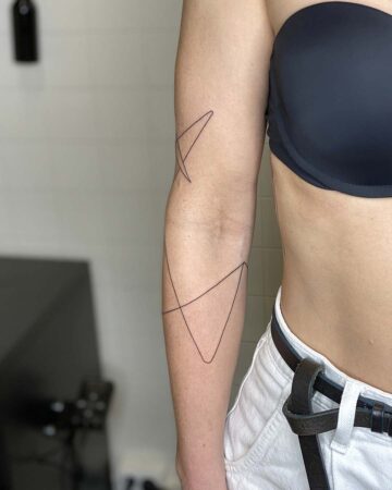 A person's arm with a minimalist line art tattoo featuring intersecting geometric shapes. The black tattoo is a simple design on the inner arm, showcasing the clean lines of a geometric tattoo. The person wears a black bra and white pants. Done by tattoo artists in our fine line tattoo studio parlour shop Canggu, Bali