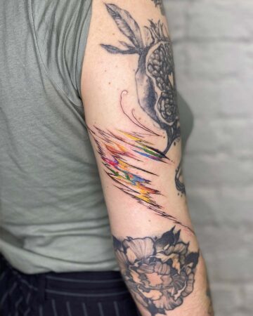 A person's arm showcases a vibrant, abstract colorful tattoo with black lines and splashes of rainbow hues. Other tattoos include a pomegranate and a large floral design. The arm tattoo is an example of modern body art. Done by tattoo artists in our fine line tattoo studio parlour shop Canggu, Bali