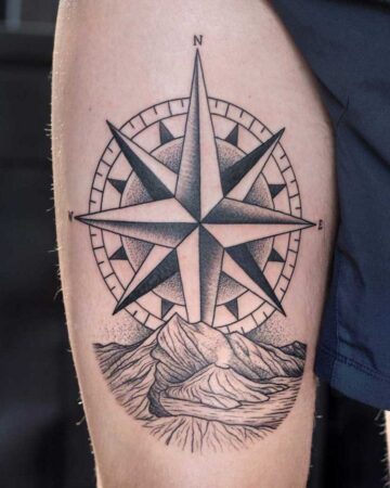 Detailed black and grey compass tattoo on a person's thigh, featuring a bold, eight-pointed star design. The compass sits above a detailed mountain range, hinting at adventure and direction. "N" and "E" are visible, indicating north and east. Done by tattoo artists in our fine line tattoo studio parlour shop Canggu, Bali