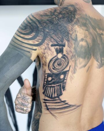 A man's back features a bold tattoo design of a train, rendered in black ink. His arm is heavily tattooed, with a thumbs-up gesture. This is a striking example of modern body art and tattoo design. Done by tattoo artists in our fine line tattoo studio parlour shop Canggu, Bali