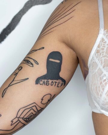 Close-up of a person's arm, showcasing a bold tattoo of a balaclava-clad figure with the word "SABOTËR" underneath, representing a unique design in body art. The arm also features other abstract tattoos, highlighting the art and design of tattoos. Done by tattoo artists in our fine line tattoo studio parlour shop Canggu, Bali