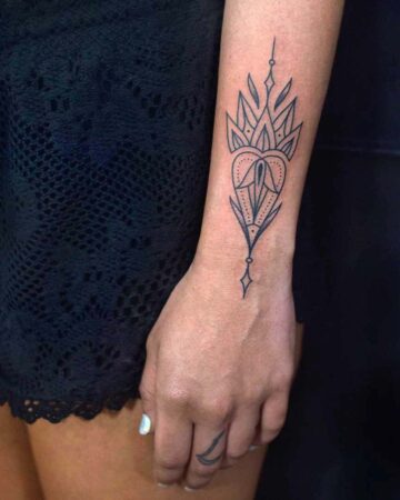 A minimalist wrist tattoo featuring a symmetrical design with geometric and floral elements. The black ink tattoo sits on the inner wrist of a woman's arm, complemented by a small finger tattoo. This wrist tattoo is a great example of fine line tattoo work. Done by tattoo artists in our fine line tattoo studio parlour shop Canggu, Bali