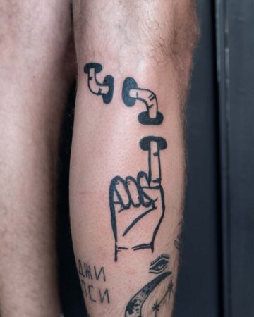 A whimsical black ink tattoo on a leg, featuring three stylized pipes and a hand making the "one" sign, with other tattoos visible below. The design is simple and bold, showcasing creative tattoo art. Done by tattoo artists in our fine line tattoo studio parlour shop Canggu, Bali