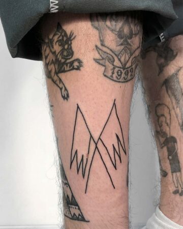 Close-up of a leg covered in various tattoos, including a simple mountain range design, a tiger, and a banner with the year 1999. The tattoos showcase diverse tattoo art and styles. Done by tattoo artists in our fine line tattoo studio parlour shop Canggu, Bali