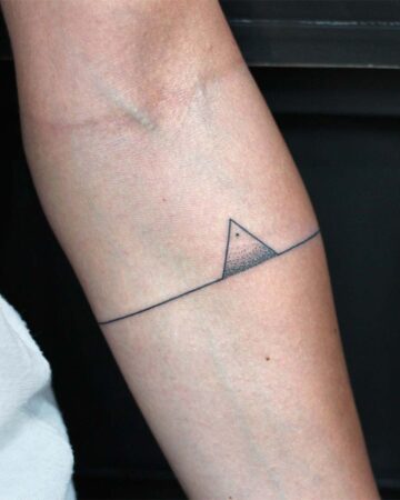 A minimalist tattoo on an arm features a thin black line with a small triangle, partially shaded with dots, above the line. The design represents a simple mountain range or landscape, popular in minimalist tattoo designs. Done by tattoo artists in our fine line tattoo studio parlour shop Canggu, Bali