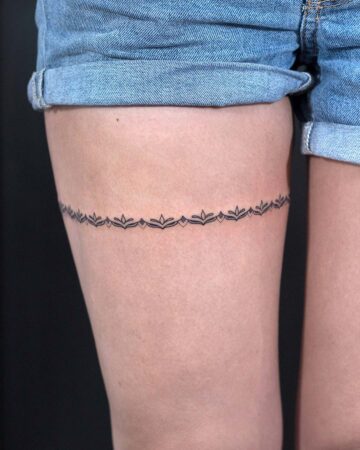 A close-up of a woman's thigh, adorned with a delicate, black garter thigh tattoo. The minimalist design features repeating floral and leaf motifs, creating an elegant, subtle look. The woman wears denim shorts, with the tattoo artfully placed just below the shorts' hemline. This is a beautiful example of a thigh tattoo. Done by tattoo artists in our fine line tattoo studio parlour shop Canggu, Bali