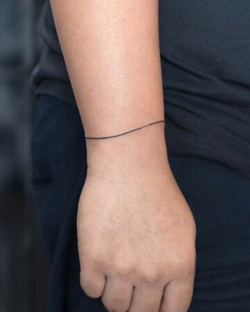 Close-up of a person's wrist with a delicate, minimalist tattoo. A thin, black line wraps around the wrist, with three tiny dots near the top. A simple and elegant design, showcasing a fine line tattoo. Perfect for minimalist tattoos. Done by tattoo artists in our fine line tattoo studio parlour shop Canggu, Bali