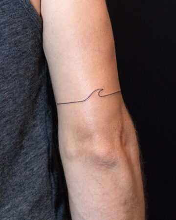 Close-up of an arm featuring a minimalist wave tattoo design. The black ink wave tattoo is a simple line drawing, a popular minimalist tattoo style. The arm is positioned against a black background. Done by tattoo artists in our fine line tattoo studio parlour shop Canggu, Bali