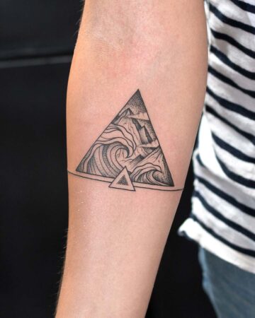 A black ink triangle tattoo on a forearm features a mountain and wave design. A small triangle sits below the main design, with a thin line connecting the two. The minimalist tattoo is a great example of a geometric tattoo. Done by tattoo artists in our fine line tattoo studio parlour shop Canggu, Bali