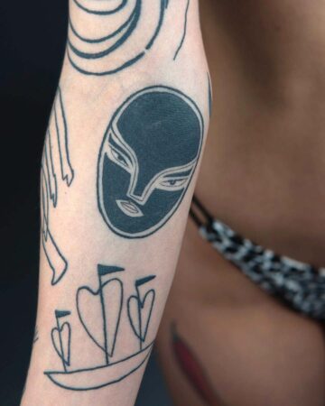 Close-up of an arm featuring several minimalist tattoos: a dark filled-in mask, a boat with heart-shaped sails, and abstract lines. The tattoo design is a modern example of body art. Done by tattoo artists in our fine line tattoo studio parlour shop Canggu, Bali