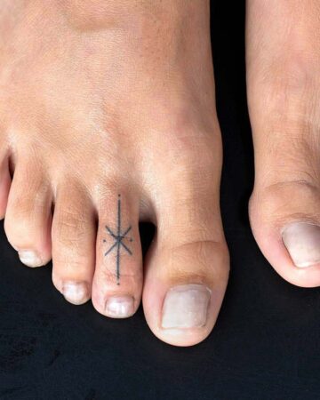 Close-up of a foot with a minimalist tattoo on the second toe. The tiny tattoo design is a simple starburst shape, a popular small tattoo idea. The foot is on a black surface, showcasing the foot tattoo. Done by tattoo artists in our fine line tattoo studio parlour shop Canggu, Bali